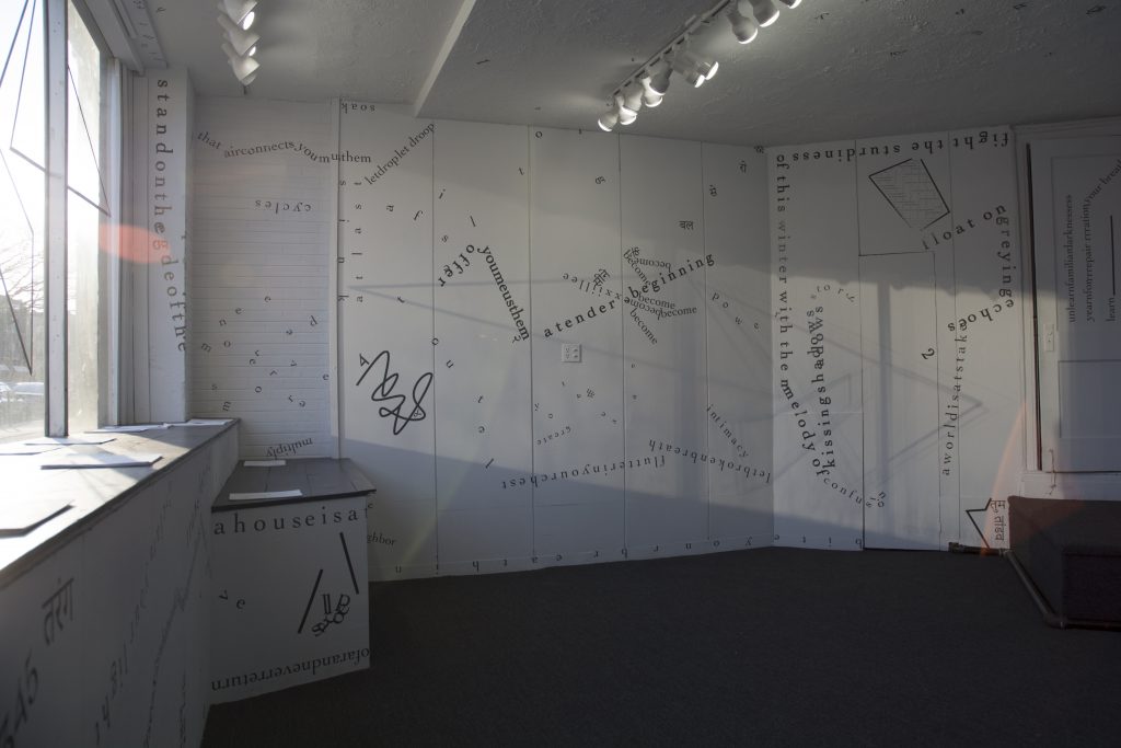 The photograph shows one full wall and parts of others as well as the front window of the gallery, with no people present. Light from the window casts shadows across the walls. Black vinyl letters are installed directly onto the white gallery walls, in the form of words and phrases in English and Hindi. Text appears in different sizes and spatial orientations (e.g., right-side up, upside-down, diagonal, vertical, and organic shapes), with some words/phrases expanded in space, condensed, or intersecting with other text. Gestural drawings—also made of black—are shown on the left-hand side of the image.