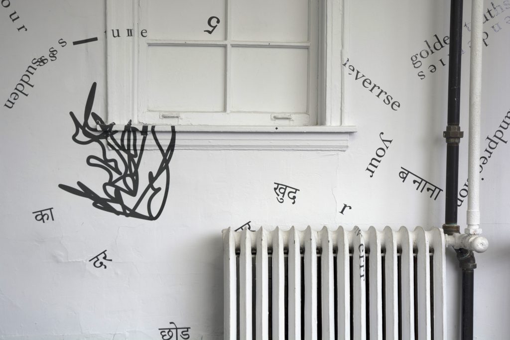 The photograph shows part of one of the gallery’s internal, white walls, including part of a window (with white glass) and radiator, and some piping. Black vinyl letters are installed directly onto these surfaces and the walls, in the form of words and phrases in English and Hindi. Text appears in different sizes and spatial orientations (e.g., right-side up, upside-down, diagonal, vertical, and organic shapes), with some words/phrases expanded in space, condensed, or intersecting with other text. English words/phrases shown in this image include “5,” “your,” and “sudden.” A gestural drawing—also made of black vinyl—is shown on the left-hand side of the image.