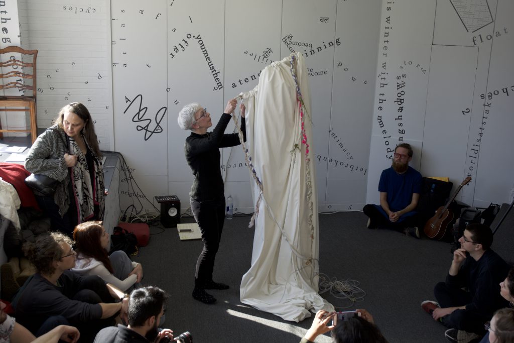 This is a still image of a performance. The performers stand in the middle of the floor, with one covered (not directly visible) in a white cloth from which a couple of colorful, braided cloths hang like ribbons or ropes. The other performer pulls or arranges one of the ribbons or ropes. Audience members are all around, leaning against a gallery wall or sitting or standing on the floor. Words, phrases, and shapes on the gallery wall and front window are visible in the background.