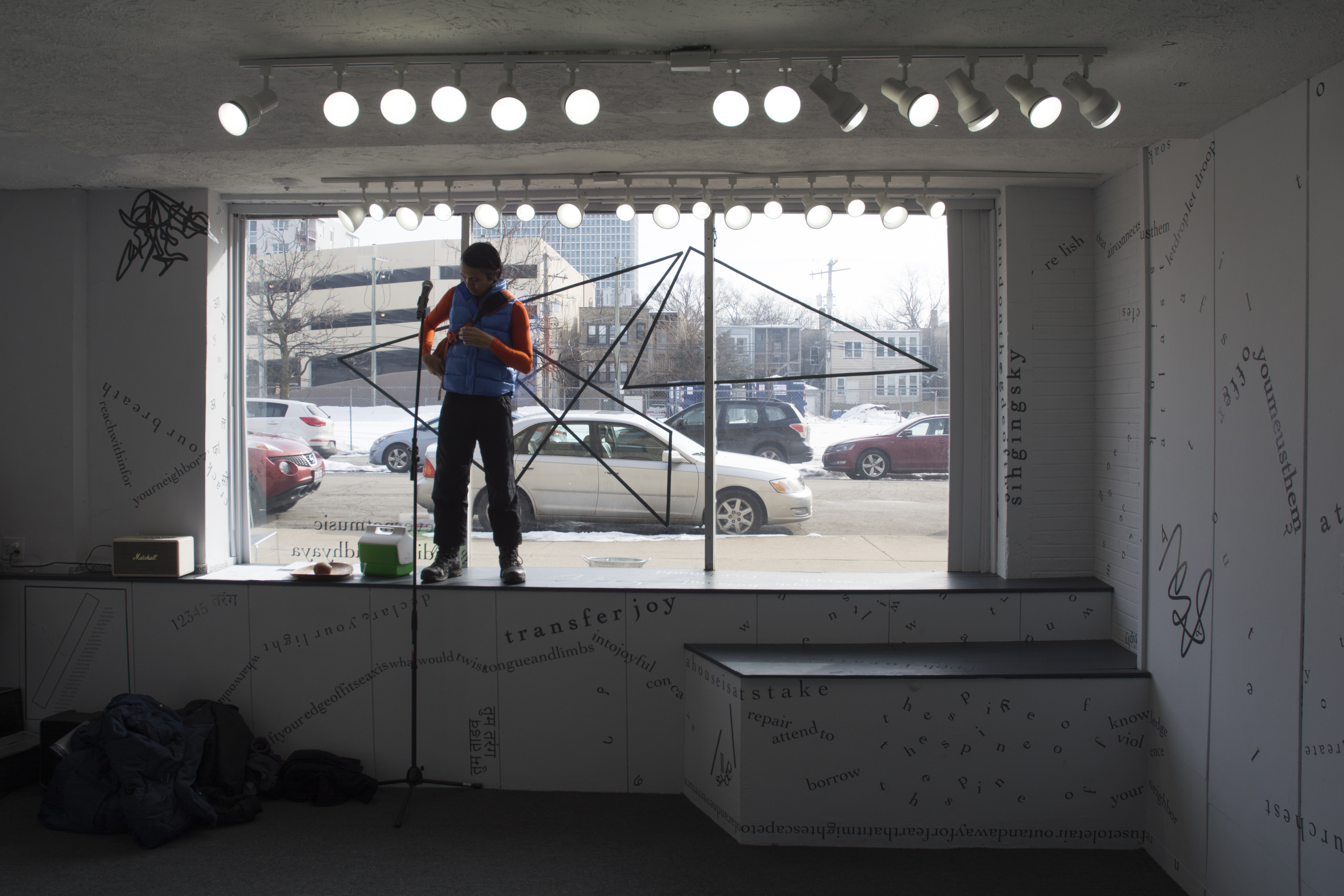 The photograph is a long shot of the inside of the gallery, with the large front window in the background and, beyond that, cars and buildings outside. A performer stands in the windowsill. Around the performer, on the sill or the floor, are a thermos, a microphone on a long stand, and a pile of cloth. Above the performer is a line of track lights that face the camera, in addition to the daylight coming in from outside. Black vinyl letters are installed directly onto the white gallery walls, in the form of words and phrases in English and Hindi. Text appears in different sizes and spatial orientations (e.g., right-side up, upside-down, diagonal, vertical, and organic shapes), with some words/phrases expanded in space, condensed, or intersecting with other text. Gestural drawings—also made of black—are shown on the top left-hand side of the image.