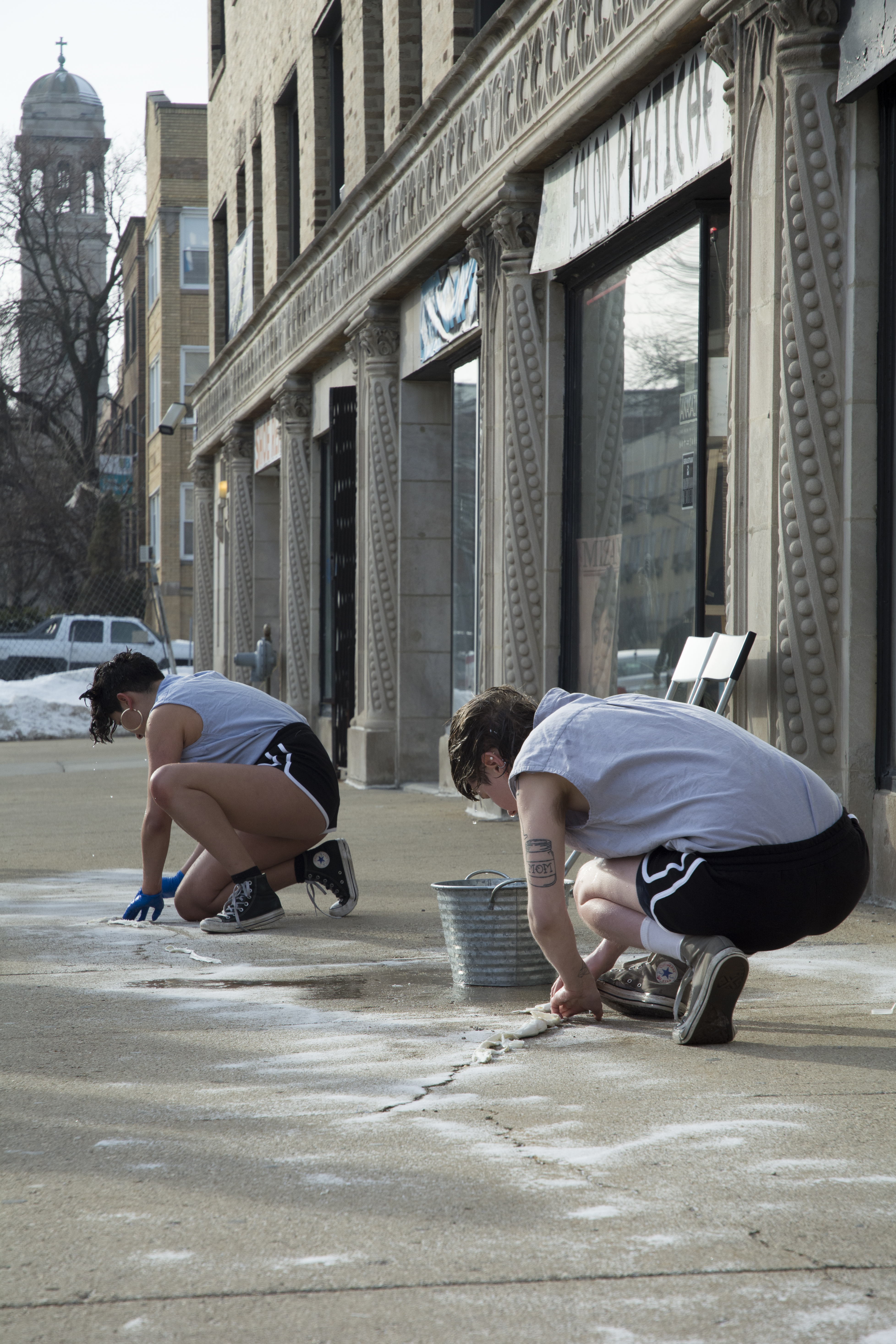 This is a still image of a performance taking place on the sidewalk outside the gallery. Two performers in matching outfits (grey-blue sleeveless shirt, black athletic shorts with white stripes, Converse All-Star sneakers) crouch on the sidewalk, facing away from the camera. A metal basin and puddle of water are on the sidewalk between them, and their hair is wet and dripping. Their hands are on the ground (one performer wearing blue gloves and the other not wearing gloves), pressing or stretching a white cloth or paper onto the concrete. Behind them are storefronts, buildings, and a sizable pile of snow.