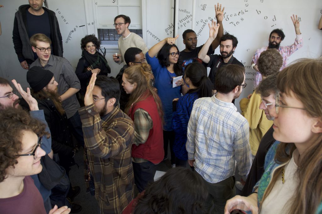 Approximately 20 people circulate inside the gallery, some close to the camera and some farther away. They appear to be moving in different directions than each other and several are raising their hands in the air. Many are smiling or laughing. 