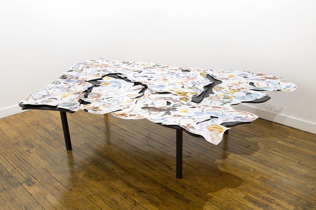 This is a photo of a corner of a gallery space, with a wood floor and with two white walls meeting in the background. In the center is a multi-layered artwork spread across a large black table. The table is asymmetrical, with many curved edges. On the table are several pieces of white paper (also asymmetrical, with many curved edges) covered in gestural drawings of various colors.