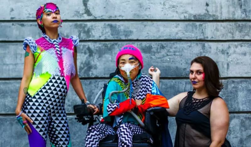 This image shows three people posing in front of a concrete wall. The person on the left is standing, at center, seated in a motorized wheelchair and wearing a breathing apparatus, and at right, kneeling. All are wearing brightly colored clothing with bold patterns.