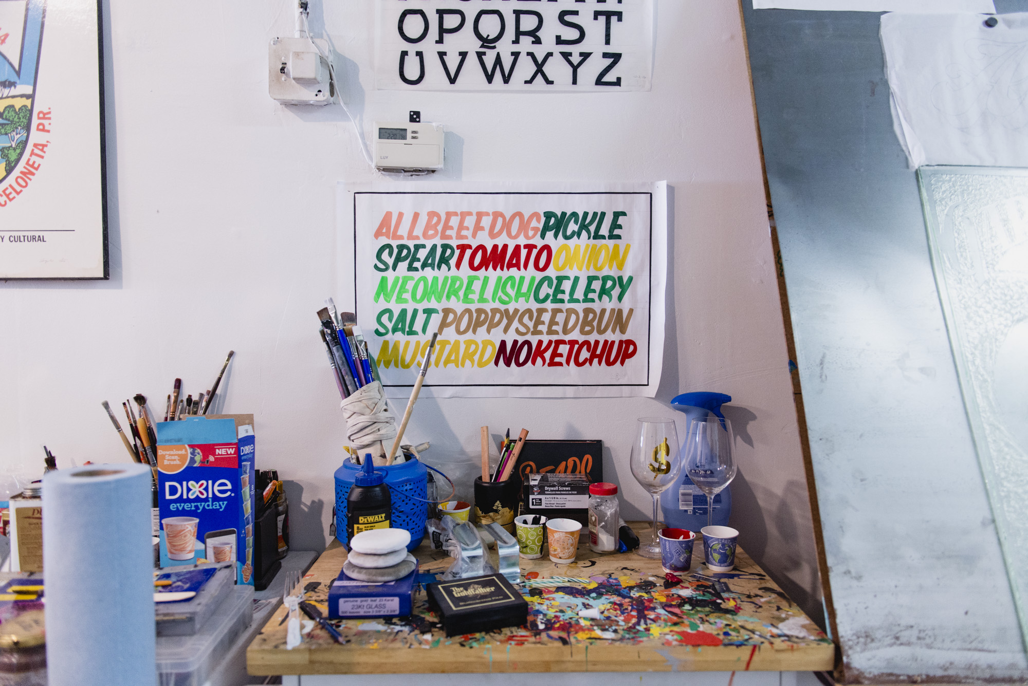 Image: Close up of various materials and tools used by Kelsey and Andrew. There are signs on the wall and colorful paint on the table. Photo by Ryan Edmund Thiel.