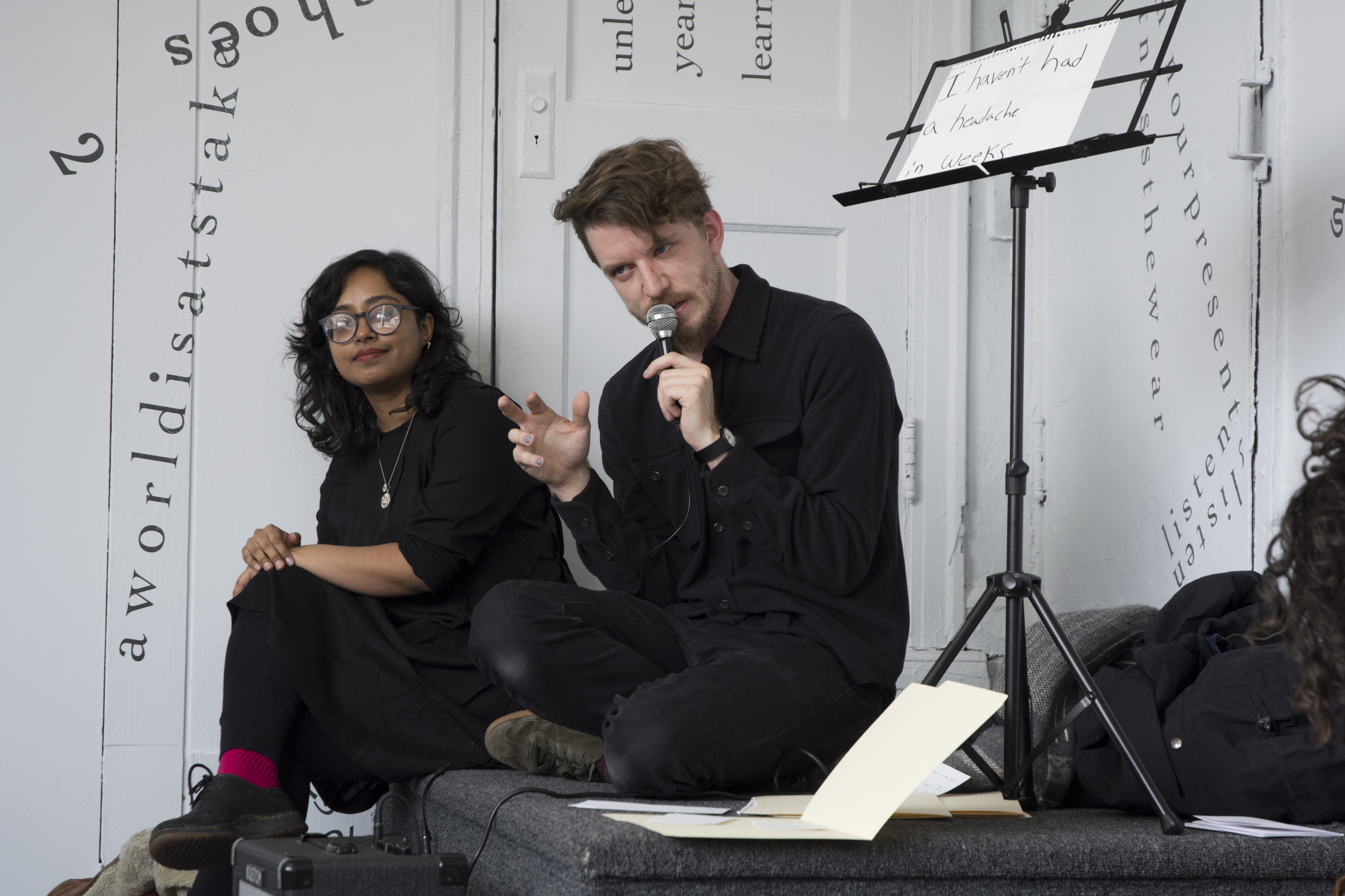 The performer and artist sit on a step in one of the gallery’s corners. Ethan speaks into a microphone while gesturing with the other hand. Above Ethan, on the right side of the frame, is a music stand holding a piece of notebook paper that reads, “I haven’t had a headache in weeks” (handwritten in black marker). Udita sits on the left side of the frame, looking toward the camera; her glasses reflect the light. Behind them are white walls and a white door onto which black vinyl letters are directly installed, in the form of words and phrases in English (including “aworldisatstake” and “listen”). Text appears in different sizes and spatial orientations (e.g., right-side up, upside-down, diagonal, vertical, and organic shapes), with some words/phrases expanded in space, condensed, or intersecting with other text.