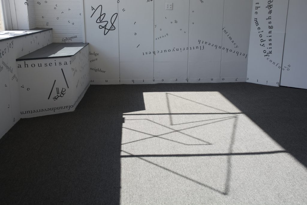 This photo shows part of the floor of the gallery, with sunlight from outside casting the shadow of the windowsill, the windowframe, and a motif of intersecting vinyl triangular shapes onto the grey rug. Black vinyl letters are installed directly onto the white gallery walls, in the form of words and phrases in English and Hindi. Text appears in different sizes and spatial orientations (e.g., right-side up, upside-down, diagonal, vertical, and organic shapes), with some words/phrases expanded in space, condensed, or intersecting with other text. Gestural drawings — also made of black — are shown on the top left-hand side of the image.