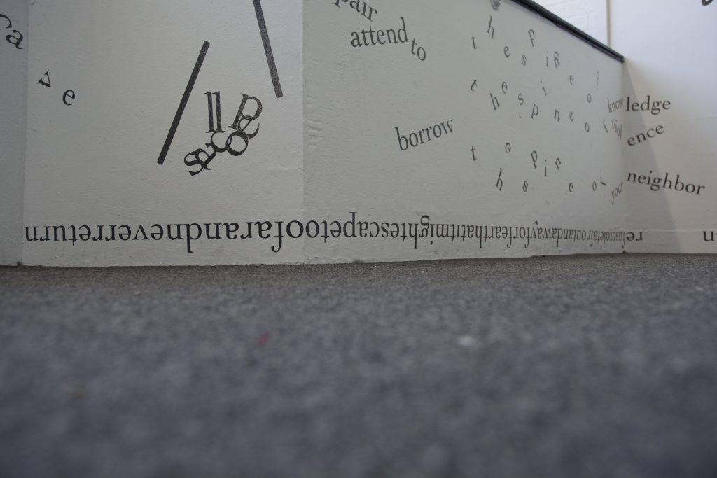 The photograph is shot from floor level and shows a low section of white gallery wall and the grey rug below. On the wall, black vinyl letters are installed directly onto the white surfaces, in the form of words and phrases in English. Text appears in different sizes and spatial orientations (e.g., right-side up, upside-down, diagonal, vertical, and organic shapes), with some words/phrases expanded in space, condensed, or intersecting with other text. A line of uninterrupted text with no space breaks runs upsidedown directly above the floor, around a convex corner.