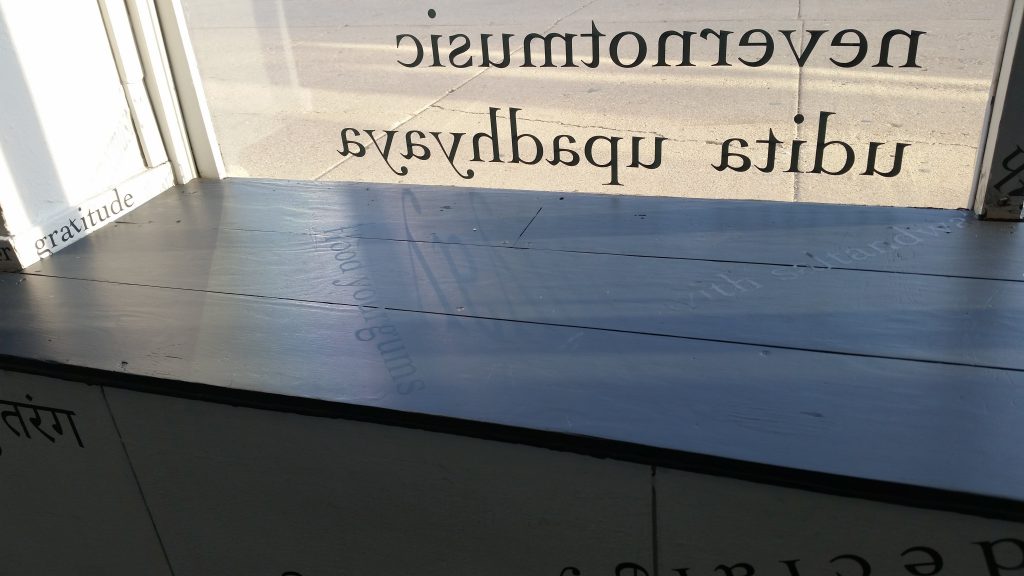 Through the glass (at the top of the image), the words “nevernotmusic” and “udita upadhyaya” are visible in reverse from behind. The sidewalk is on the other side of the glass. There is black vinyl lettering on the black windowsill (including “flood your gums”), on the white windowframe (“gratitude”), and the white gallery wall below (in Hindi).