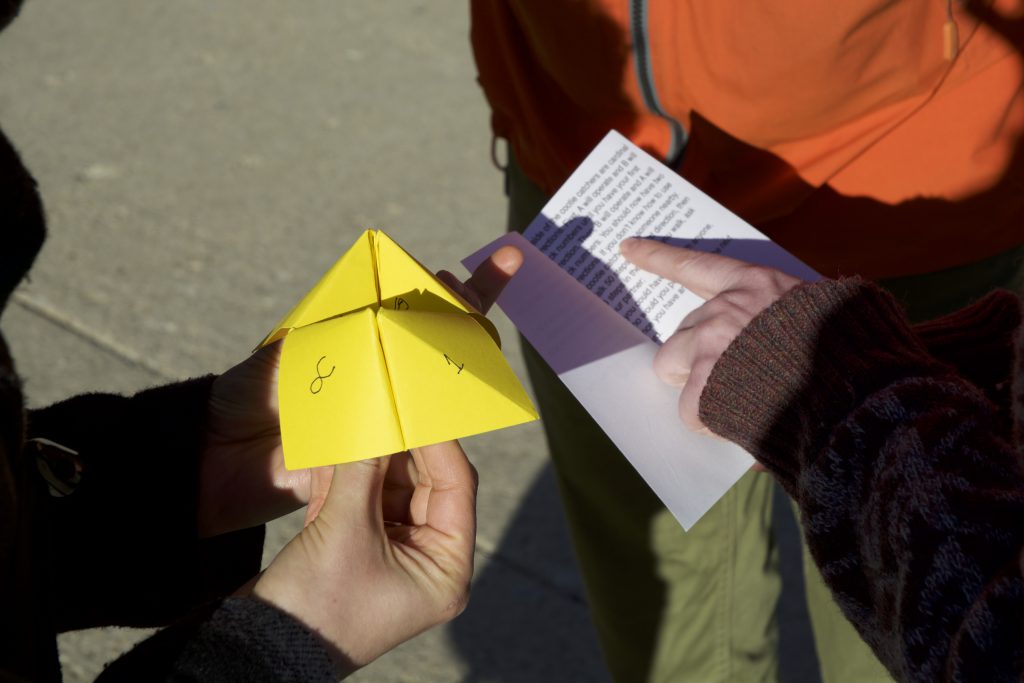 An extreme close-up of three people outside, using a cootie catcher. At left, a pair of hands holds a yellow cootie catcher. At right, another person holds a small booklet and points to its typed content. A third person stands just beyond that.