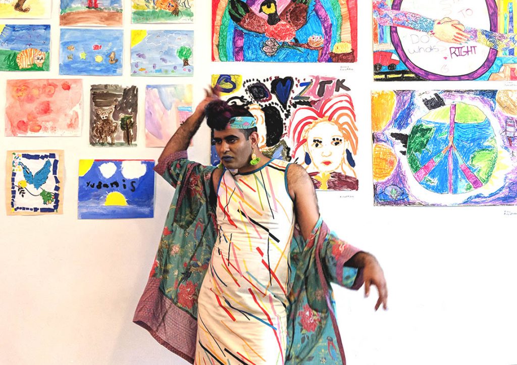 Image: Alok Vaid-Menon poses in front of a wall of colorful artwork at AMFM gallery in Pilsen before their show. They are looking away from the camera, standing statuesque with one arm lightly rested on their front top bun, hand facing up, and their other arm casually extended, in soft motion, like a queen expressing a need. Photo by Chelsea Ross.