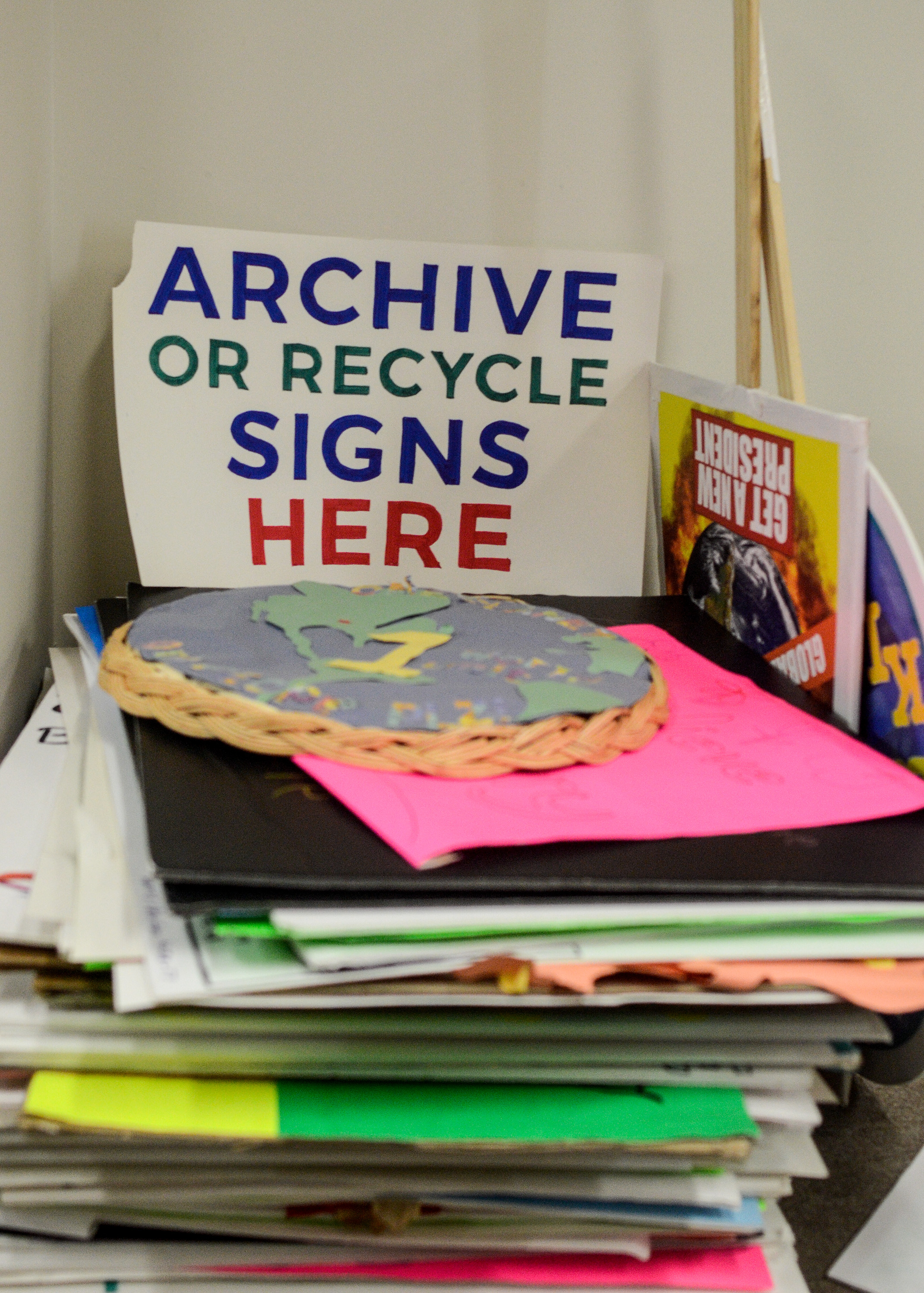 Image: A stack of multicolored poster board signs is shown. A sign that reads "Archive or Recycle Signs Here" stands above the stack. One sign is the map of the world pasted onto a woven basket. Two signs with sticks attached can be seen to the right. Image by William Camargo. 