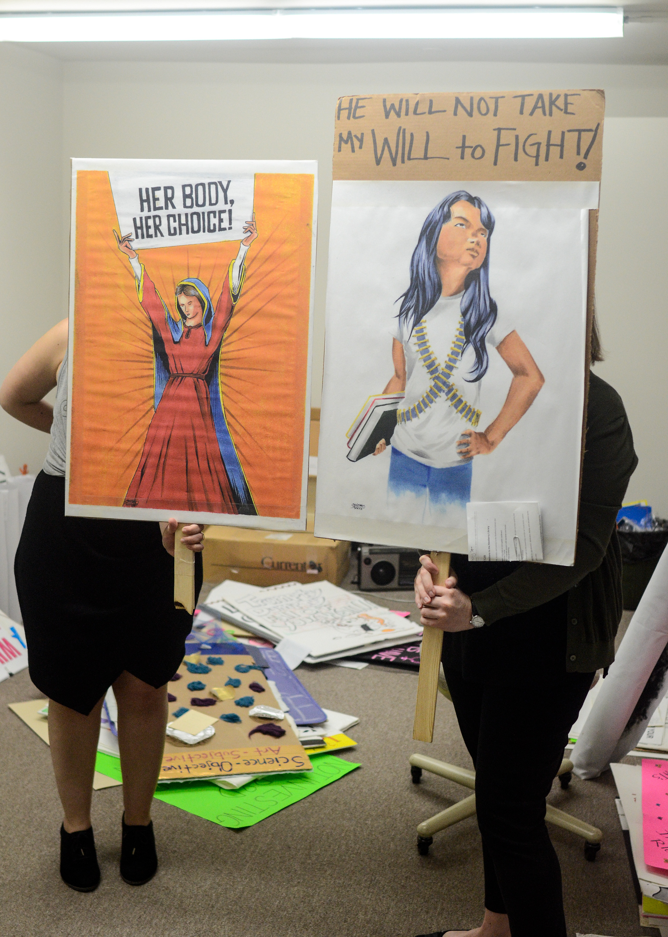 Image: Catherine Grandgeorge and Jennifer Patiño Cervantes hold up signs in front of them. The two signs are of images made by and donated by artists Adrian Santiago-Alvarez. One shows a Virgin Mary like figure holding a sign that says "Her Body Her Choice." The other shows a defiant young woman holding books and wearing bandoliers under a sign that says "He Will Not Take My Will to Fight!" Image by William Camargo. 