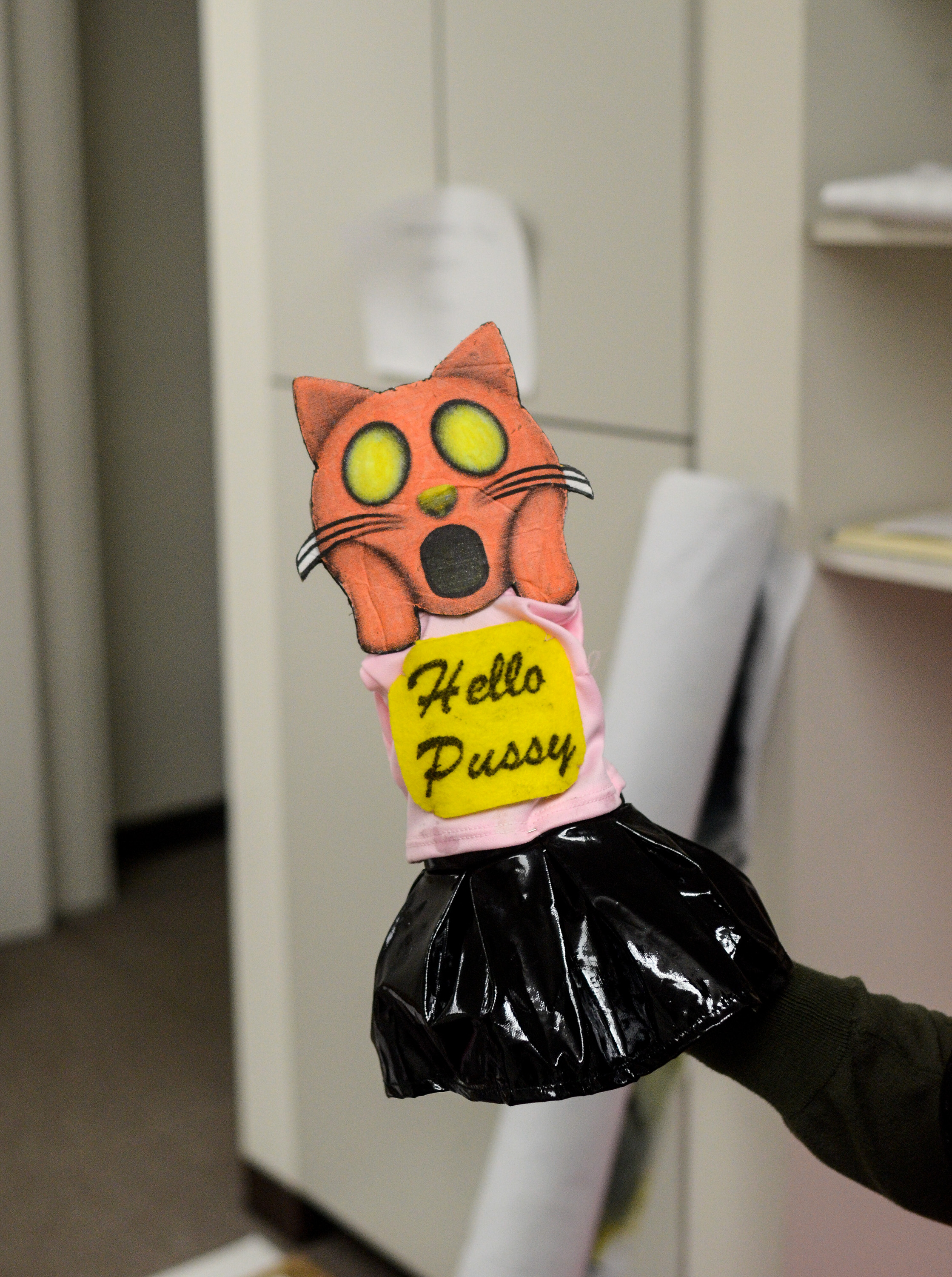 Image: A hand puppet has the head of an orange emoji cat making a shocked face that alludes to "The Scream" by Edvard Munch. It wears a pink and black outfit with a yellow sign that says "Hello Pussy." Image by William Camargo. 