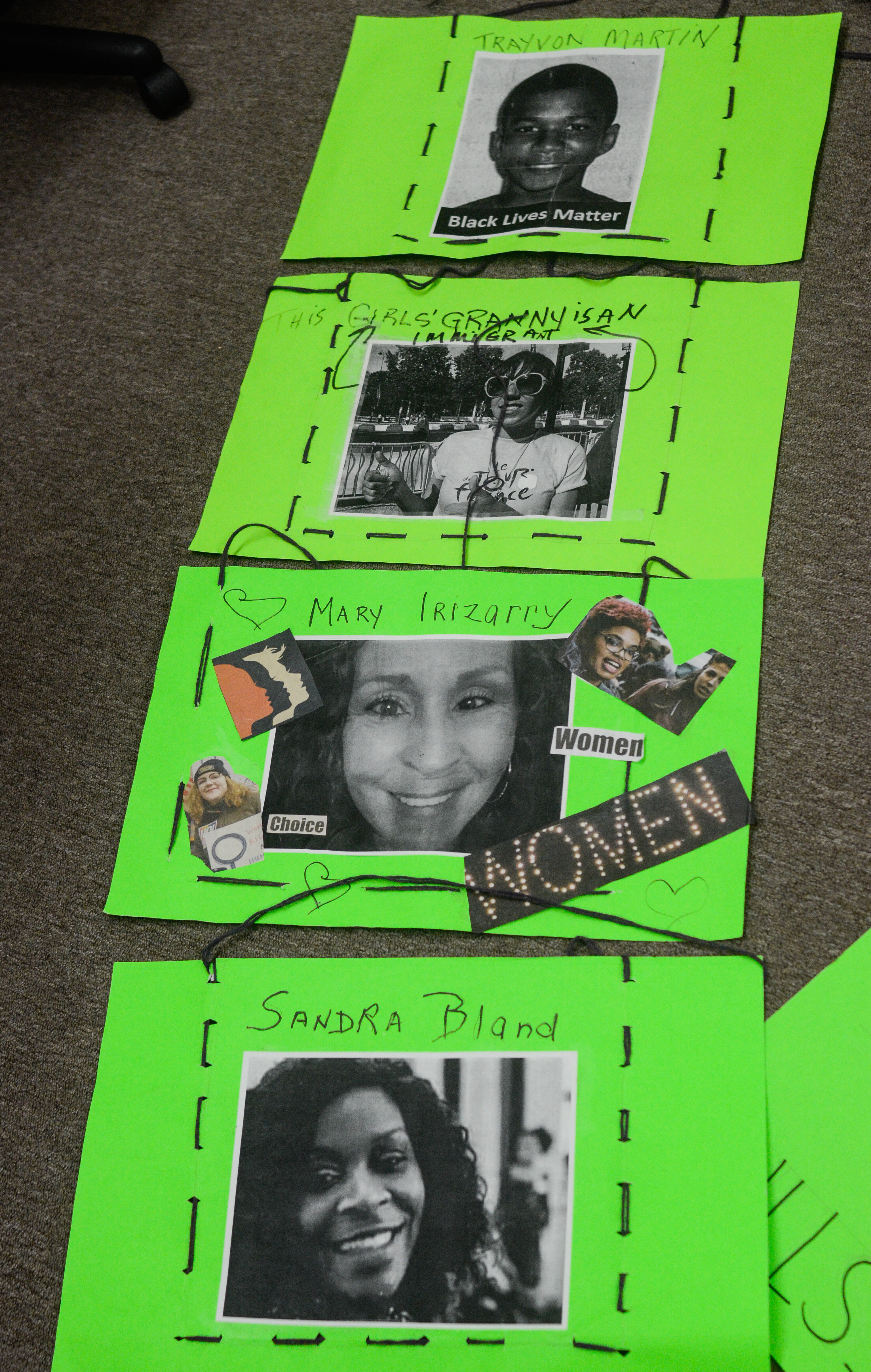 Image: Four green poster boards are stitched together with black thread. It appears designed to be draped across the front and back of the body. Photos are accompanied with text on each sign. From top to bottom, the text reads "Trayvon Martin," "This Girls' Granny is an Immigrant," "Mary Irizarry," along with "Women" and "Choice," and lastly "Sandra Bland." Image by William Camargo. 