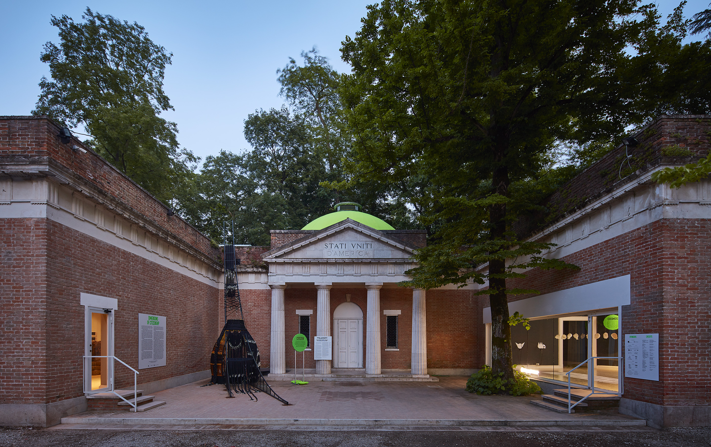 Image: The 2018 U.S. Pavilion. The image shows an exterior view of the U.S. Pavilion at dusk with light glowing from the windows of the gallery spaces. Photo © Tom Harris. Courtesy of the School of the Art Institute of Chicago and the University of Chicago.