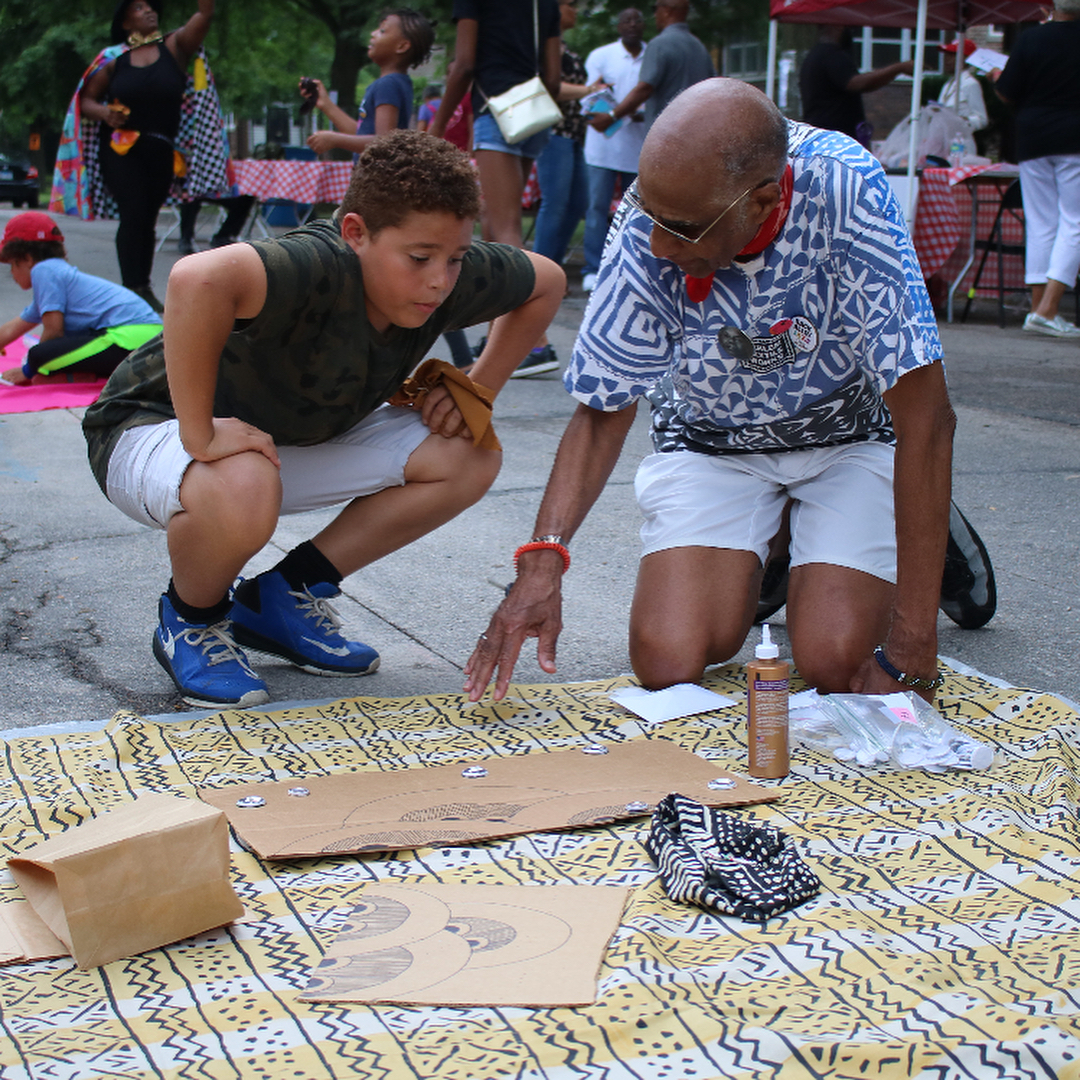 Image: A 9- or 10-year-old child with curly dark hair squats beside Robert Earl Paige as the artist gestures towards a design created on cardboard and lying flat on a textile on the concrete. Image courtesy of the Museum of Vernacular Arts.
