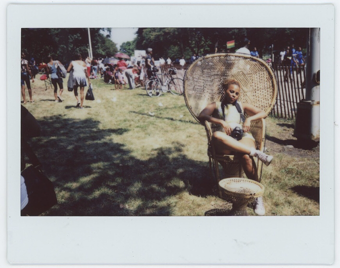 Image: Scheherezade Tillet sits in a wide-backed wicker chair on a grassy lawn full of other people, evoking Huey Newton's famous Black Panthers portrait. Image courtesy of the Museum of Vernacular Arts.