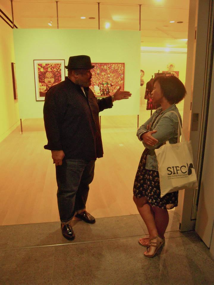 Image: Dawoud Bey and me at the Logan Center for the Arts, 2013. Photo by Ron West. An image of two people standing in the doorway of an art gallery, facing one another. The person on the left of the image is speaking, with had outstretched in front of him. The other is standing, arms and leg crossed, listening. 