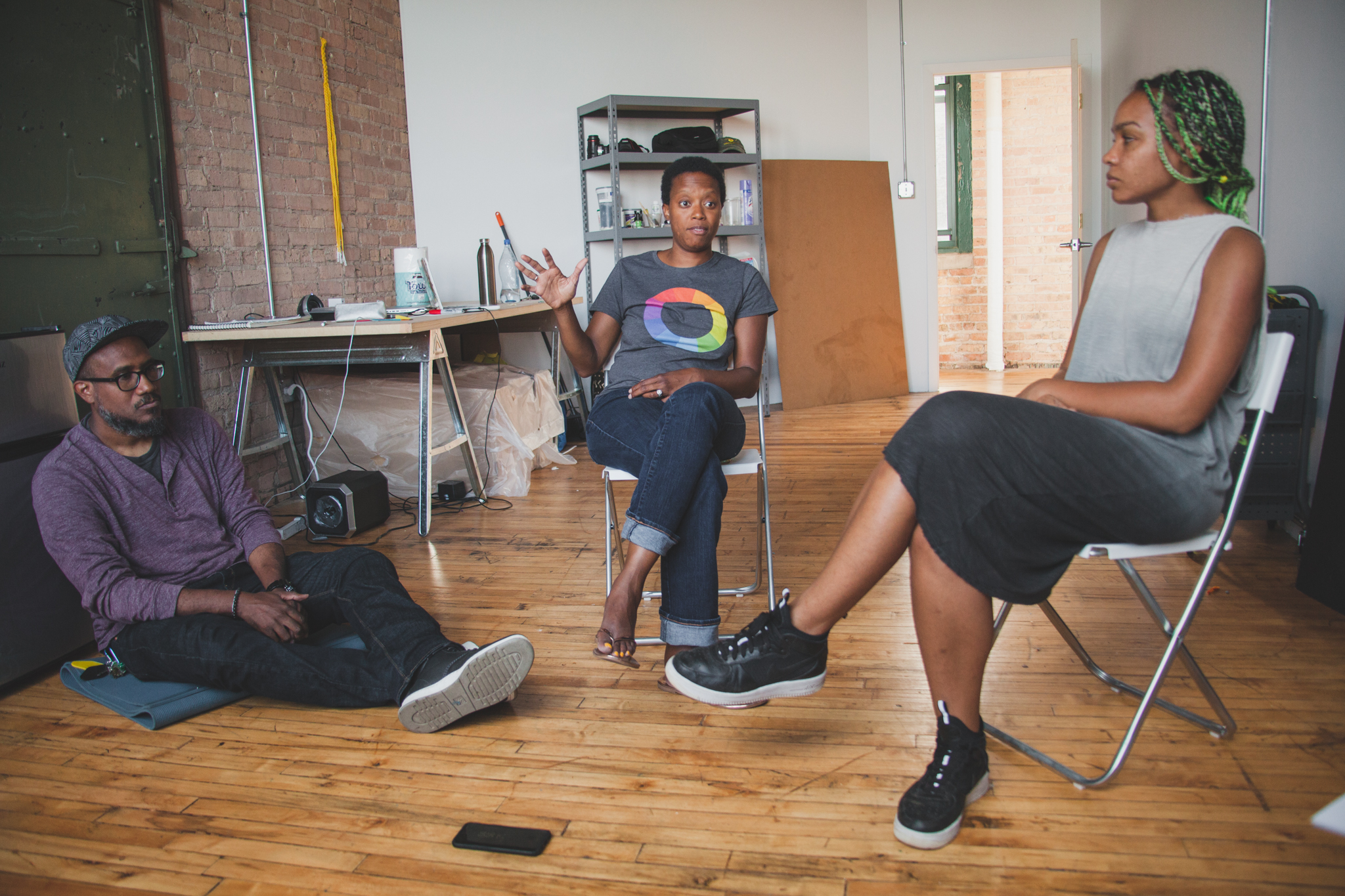 Image: Andres, Amanda, and Shani in a studio at Bridgeport Art Center, 2018. The image shows Andres sitting on the ground, Amanda and Shani sitting in chairs near him and Amanda is speaking as the other two listen. Photo by Ally Almore. 