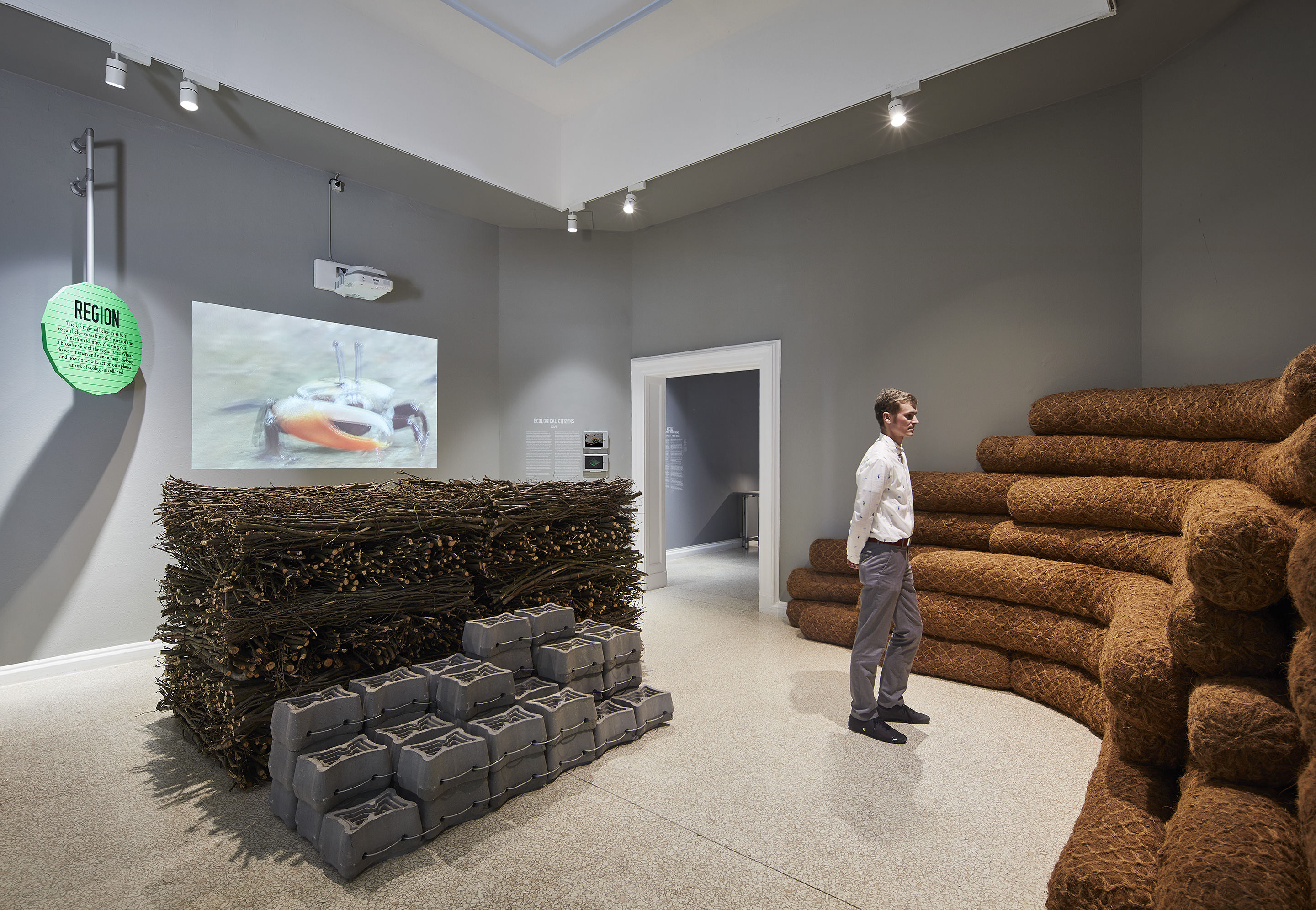 Ecological Citizens by SCAPE at the 2018 U.S. Pavilion. The image shows an installation view of an exhibition space. On the wall to the left there is a projected image. On the wall to the right there are stacks of long, brown tubes. In the center of the room is a neatly stacked cluster of wood sticks and cinderblocks. There is a person standing in the middle of the gallery space, studying the works. Photo © Tom Harris. Courtesy of the School of the Art Institute of Chicago and the University of Chicago.