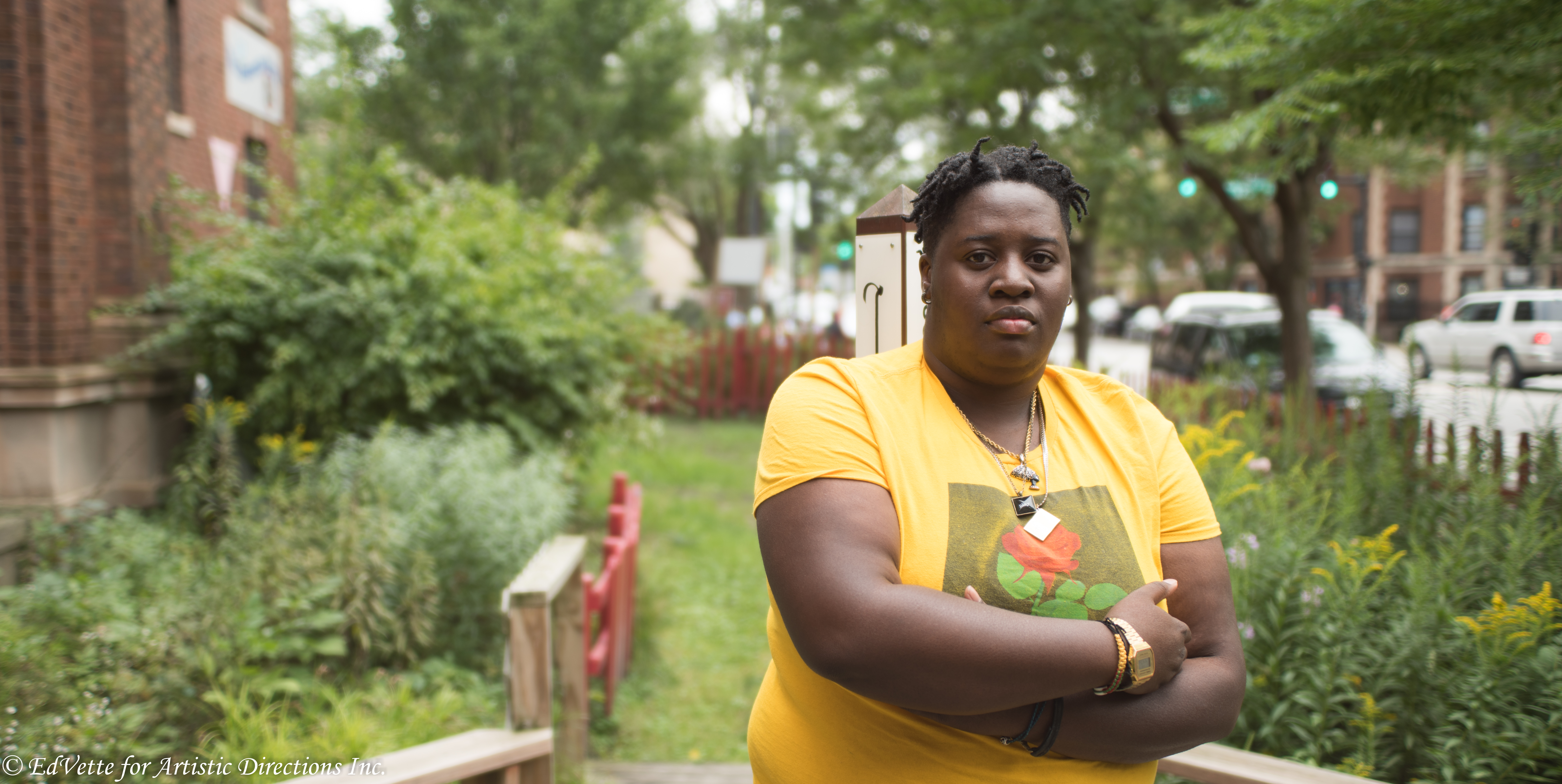 IMAGE: AnnMarie Brown stands in front of a garden area near the entrance of United Church of Rogers Park. She is staring at the camera with arms slightly folded wearing a yellow shirt with a red rose imprint on the front.