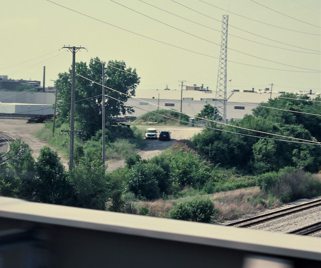 Image: On an unpaved backroad in the distance, surrounded by crisscrossing train tracks and telephone wires, two Chicago Police Department SUVs, one blue and white, one unmarked gray, sit facing opposite directions in the shade of a tree, the drivers apparently convening through their door windows. Except from this view, through the window of a passing car on the CTA Pink Line, which runs through North Lawndale, the spot is clandestine. Photo by Eric K. Roberts.