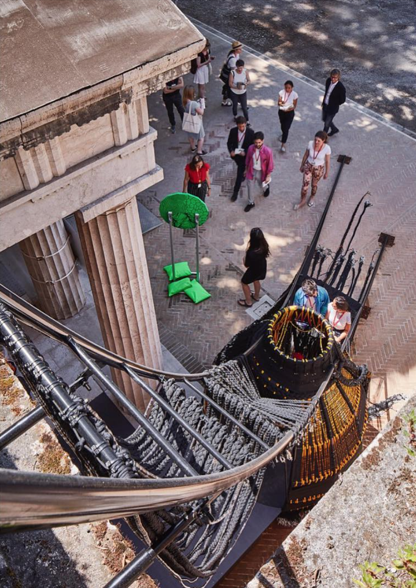 Image: Detail of Thrival Geographies (In My Mind I See a Line) by Amanda Williams + Andres L. Hernandez, in collaboration with Shani Crowe at the 2018 U.S. Pavilion. The image is from the roof of the building, looking down the structure to the ground below. The image shows a ladder-like structure swooping down to the base, and people are standing on the sidewalk near the sculpture. Photo from Shani Crowe's Instagram.