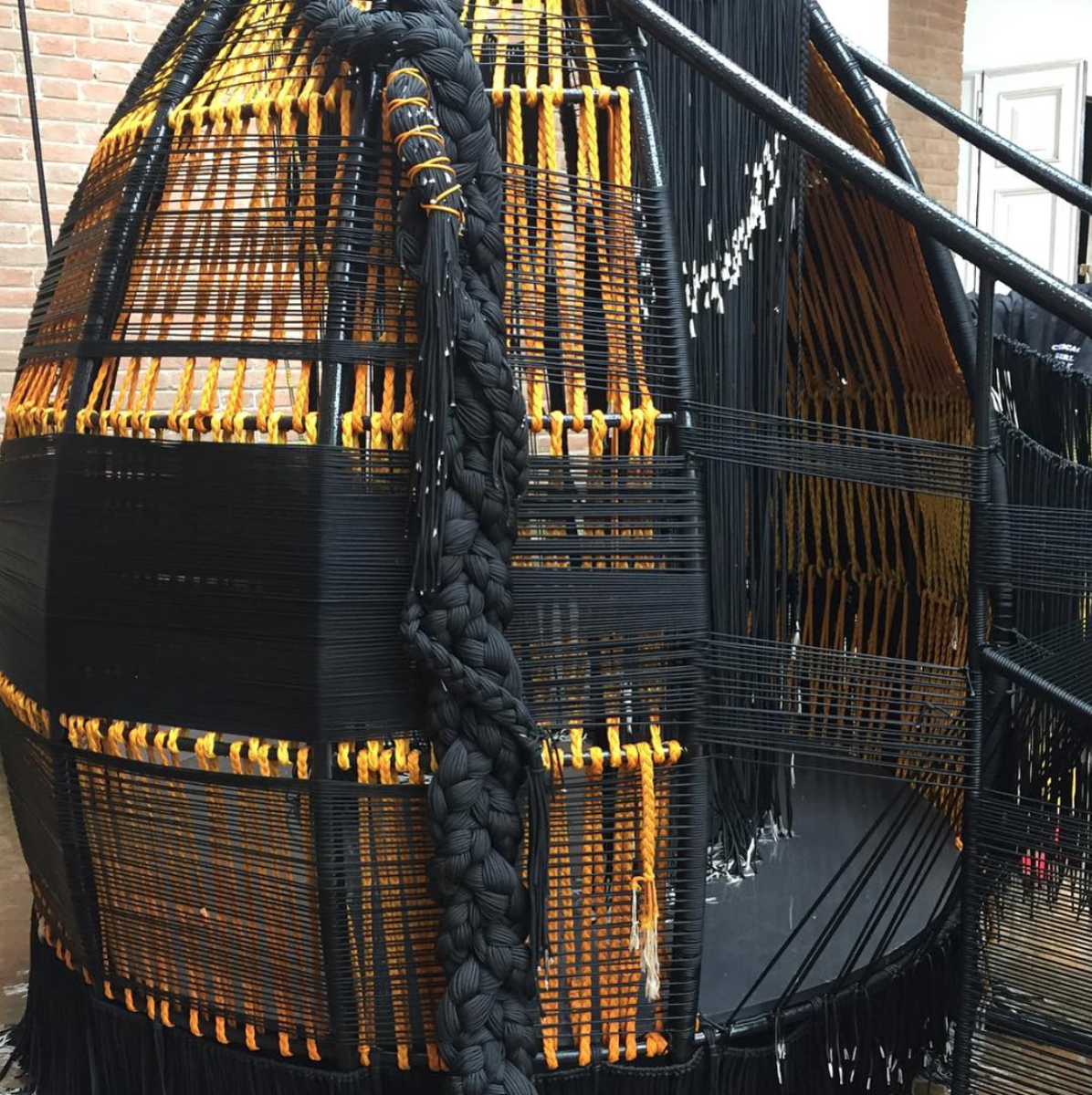 Image: Detail of Thrival Geographies (In My Mind I See a Line) by Amanda Williams + Andres L. Hernandez, in collaboration with Shani Crowe at the 2018 U.S. Pavilion. The image shows the inhabitable part of the structure, focusing on the details of the exterior, including the weaving of black and yellow ropes, and thick, black braids falling along the outer wall. Photo from Shani Crowe's Instagram.