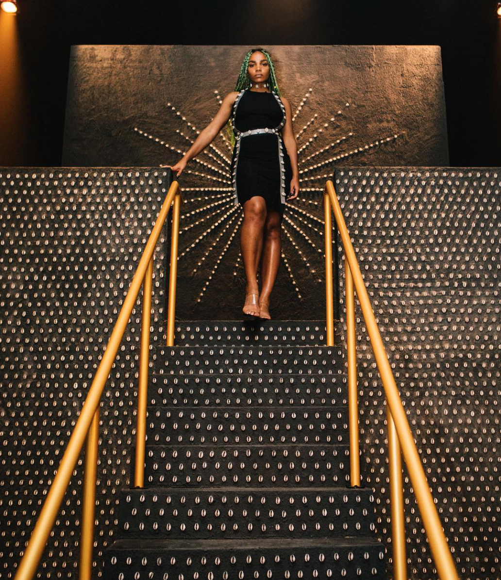 Image: Shani Crowe standing at the top of the stairs of her recent installation, Rest in Peace, at a Refinery29 event in Chicago. Behind her is a starburst made of cowry, adhered to the wall. In the foreground are the stairs and the facade of the structure, which are all patterned with cowry shells as well. The golden stair rails lead down and toward the camera. Photo from Shani Crowe's Instagram.