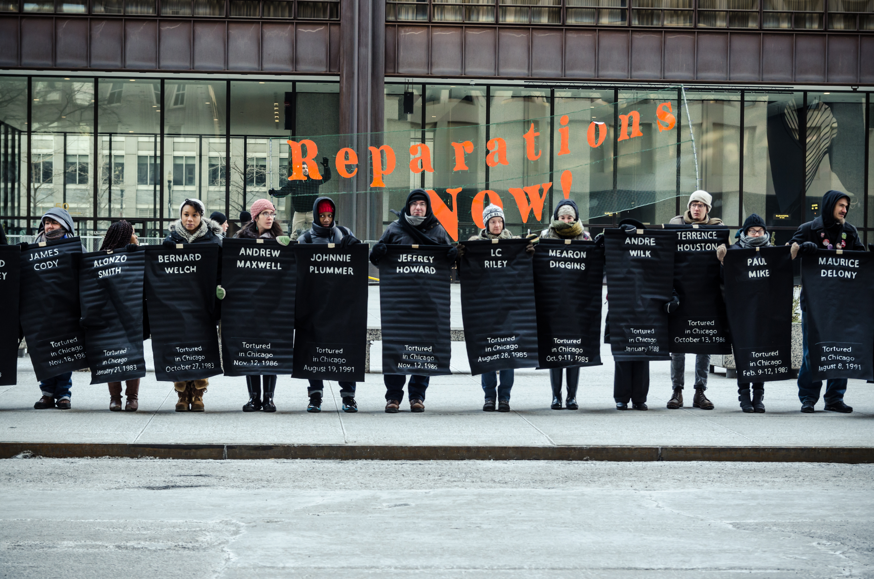 Image: People lined up facing the camera. They are holding up Black posters with the names of victims of torture by Chicago Police inscribed in white. Behind them is a transparent banner with the words “Reparations NOW!” inscribed in orange. Photo taken February 2015 at the Rally for Reparations organized by The Chicago Torture-Justice Memorials, Project NIA, Amnesty International USA, and We Charge Genocide. Photo courtesy of the artist.