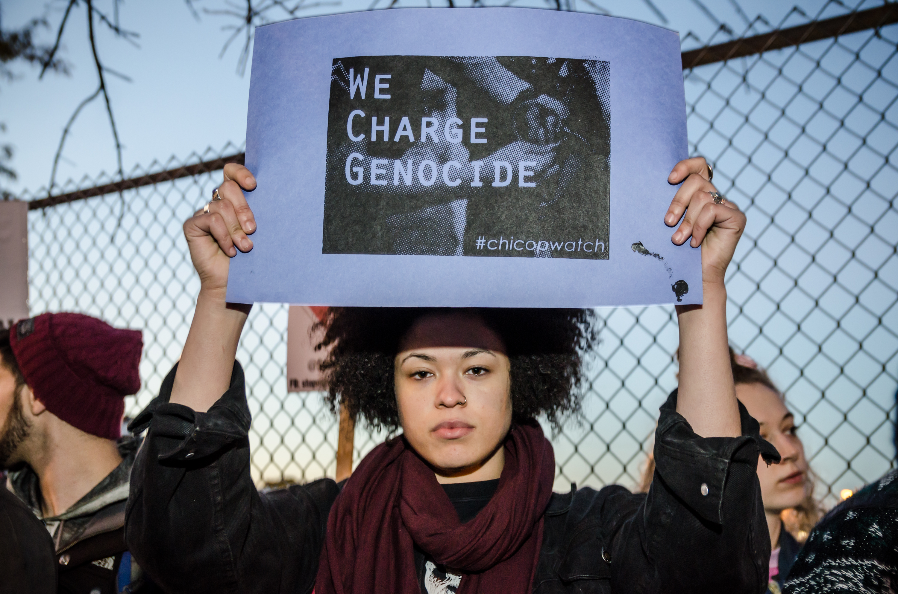 Image: A young person of color stands in front of a chain-link fence, looking directly at the camera. They are holding up a poster with an image that has the words “We Charge Genocide” super imposed in the center of the poster. The hashtag #chicopwatch is placed on the lower right corner of the poster. Photo taken October 22, 2014, at the Break Down the Wall of Silence Protest Against Police Brutality, organized by We Charge Genocide. Photo courtesy of the artist.
