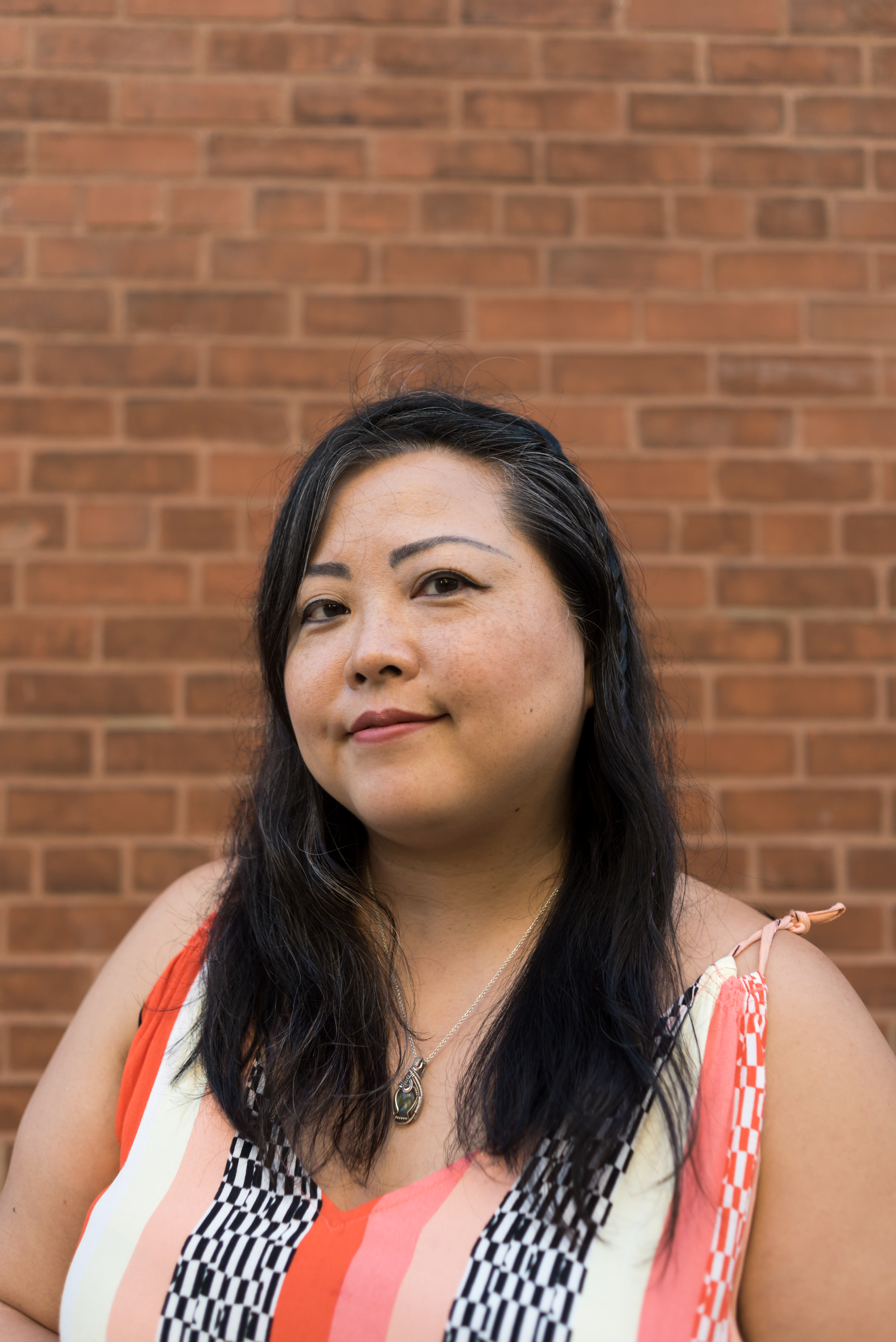 Image: A portrait of the artist Sarah-Ji standing in front of a brick wall wearing a patterned dress. She looks directly at the camera with a slight smile. Portrait taken by Ireashia Bennett.