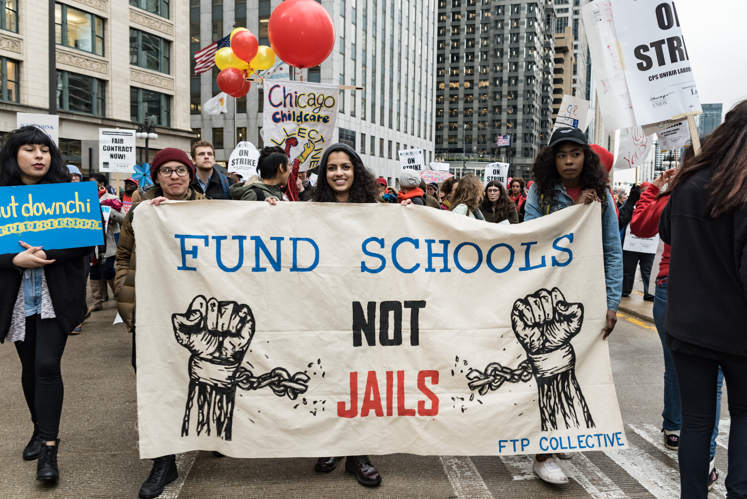 Image: Photo of a group of people rallying in downtown Chicago. In the center of the image are three people of color holding up a sign which says: “Fund Schools Not Jails” in the center. On either side of the words are images of painted fists with broken shackles around the wrists. On the bottom right of the poster is “FTP Collective.” Photo taken April 2016 at the Shut Down the Chi Rally. Photo courtesy of the artist.