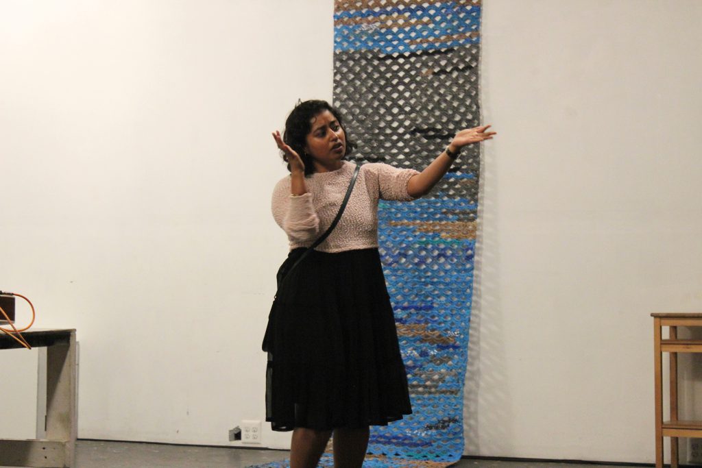 Image: Udita Upadhyaya at the “nevernotmusic” book release at TriTriangle. The artist stands at the center of the image, gesturing with both arms while speaking. Hanging on the wall behind Udita and extending onto the floor is a blue, grey, brown, and black artwork by Jerry Bleem, which was crocheted as part of a performance in response to Udita’s score, “Dear Jerry and Nick: Hold (a hand a spine a heart a whole self).” Udita wears a light-colored, textured sweater and black skirt. Photo by Caleb Neubauer.