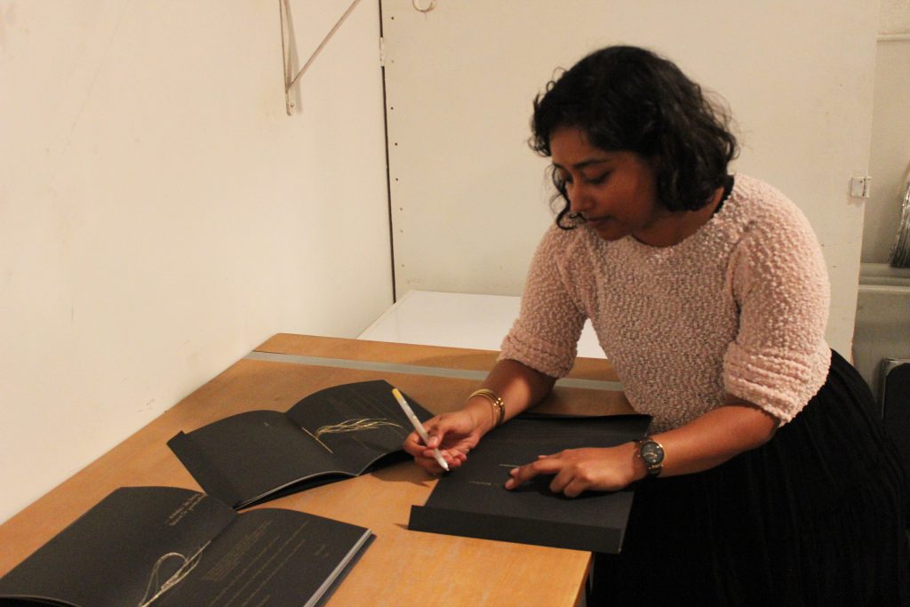 Image: Udita Upadhyaya at the book release for “nevernotmusic.” The artist leans over a table, looking down as she writes in gold pen inside a copy of her book. Next to Udita are two other copies, open to their centerfolds, where gold thread is visible. The artist wears a light-colored, textured sweater and black skirt. Photo by Caleb Neubauer.