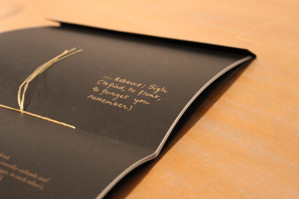 Image: Udita Upadhyaya’s book “nevernotmusic” (detail). On a wooden tabletop, the book lies open to the centerfold, showing black paper, gold thread, and gold writing. The word “Dear” is legible in gold type, followed by gold handwriting: “Rebecca: Sign (to find, to float, to forget you remember).” Photo by Caleb Neubauer.