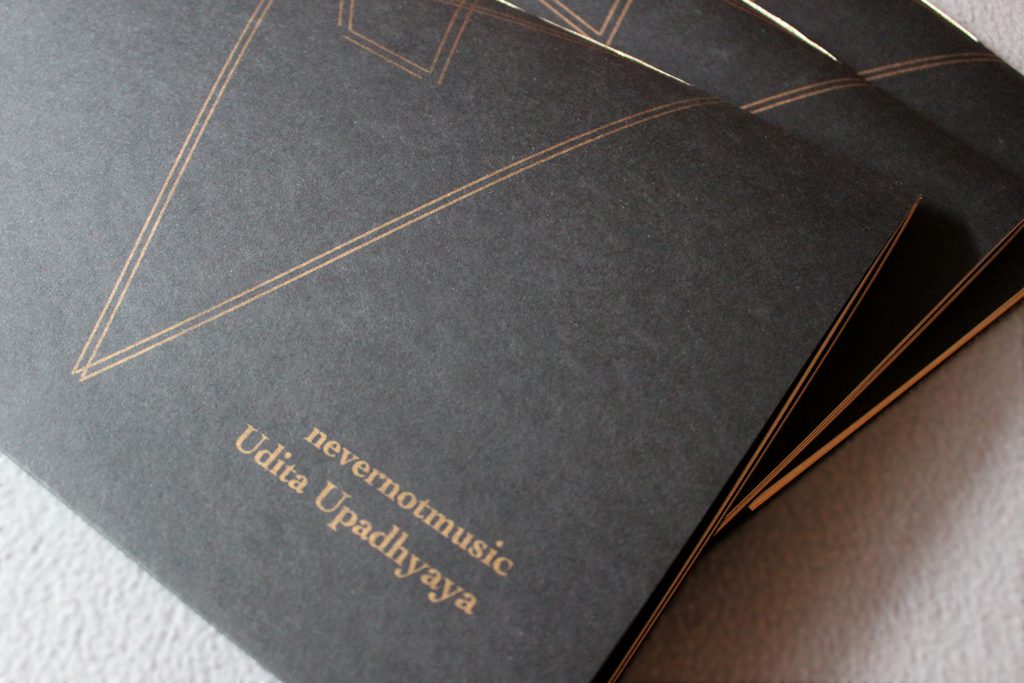 Image: Udita Upadhyaya’s book “nevernotmusic.” Three copies are fanned out on a light grey surface. Most of the front cover of the top copy is visible (showing a motif of thin, gold, parallel lines forming a large triangular and a small rhomboid shape), as is the book’s gold stitching. The cover reads “nevernotmusic” and “Udita Upadhyaya” in gold text, on black paper. Photo by Caleb Neubauer.