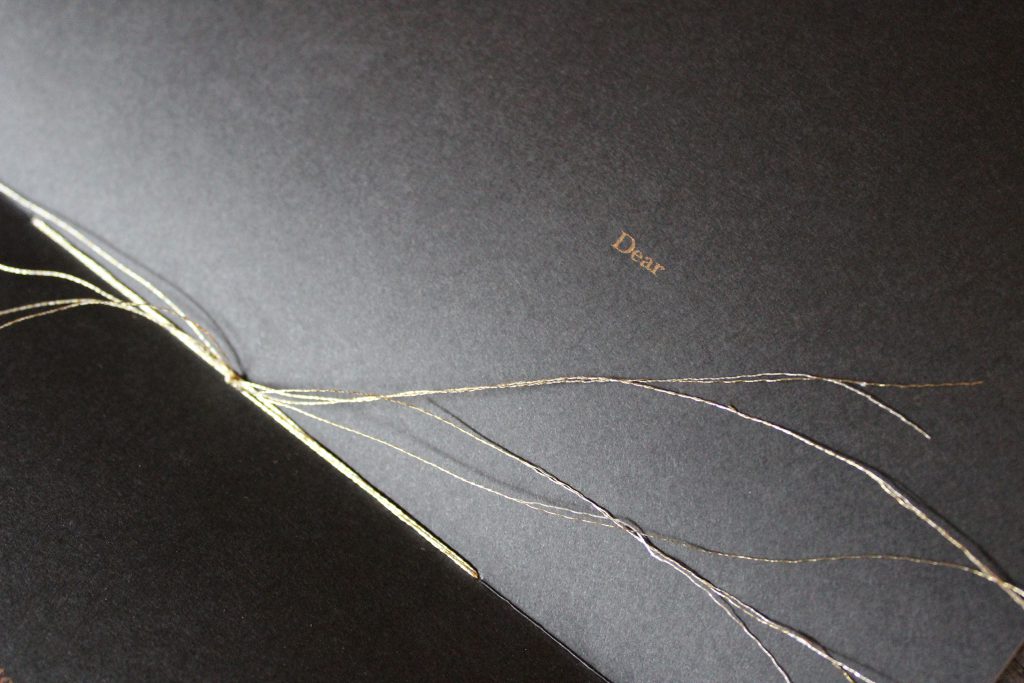 Image: Udita Upadhyaya’s book “nevernotmusic” (detail). The book lies open to the centerfold (in close-up), showing black paper, knotted gold thread (the book’s stitching), and gold type (reading “Dear”). Photo by Caleb Neubauer.