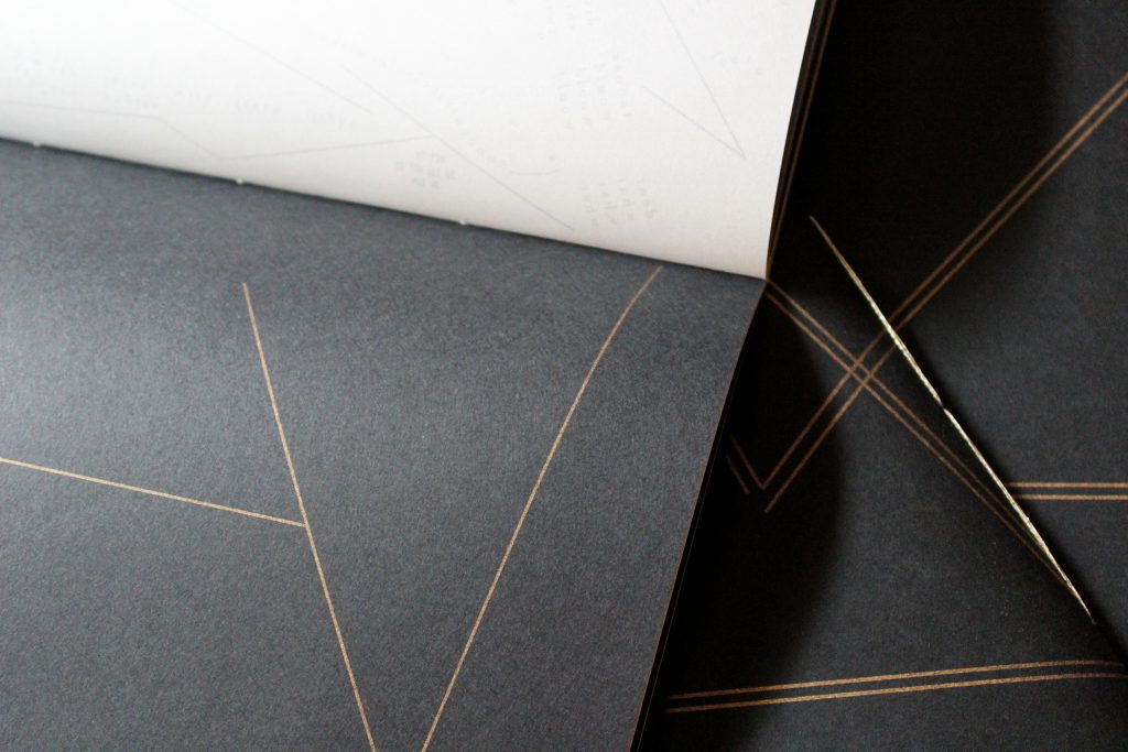 Image: Udita Upadhyaya’s book “nevernotmusic” (detail). Part of one page-spread is visible, with opaque black paper on one page (on which three thin gold lines touch to form large open triangles) and with whitish vellum on the other page (with lines, open triangles, and letters printed on the other side of the sheet of vellum, and slightly visible through it). In the background, beyond this copy, is part of the front cover of two other copies, with gold stitching and straight gold lines on black paper. Photo by Caleb Neubauer.