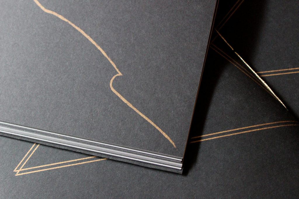 Image: Udita Upadhyaya’s book “nevernotmusic” (detail). Part of one page is visible, with a wavy, gold, gestural line (shaped somewhat like an elongated wave of water) descending across the black paper. In the background, beyond that page, is part of the front cover of two other copies, with gold stitching and straight gold lines on black paper. Photo by Caleb Neubauer.