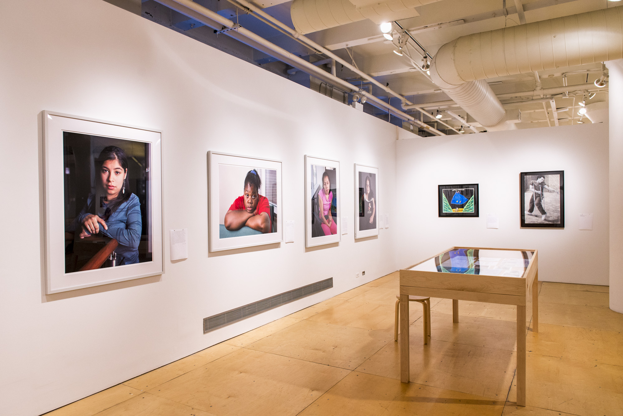 Image: a view of two gallery walls featuring primarily large format photography portraits; to the right is a display table with printed ephemera. Photo by Ryan Edmund. 