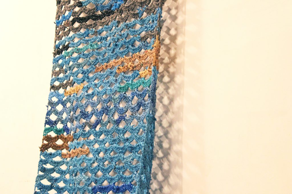 Image: Detail of a crocheted piece by Jerry Bleem, which was created as part of a performance in response to Udita’s score, “Dear Jerry and Nick: Hold (a hand a spine a heart a whole self).” The piece is made of blue, grey, brown, and black plastic bags. The crocheted pattern appears like a series of connected crescents, with the white wall visible behind the artwork and through the gaps between the crescents (in turn, casting patterned shadows on the wall). Photo by Caleb Neubauer.