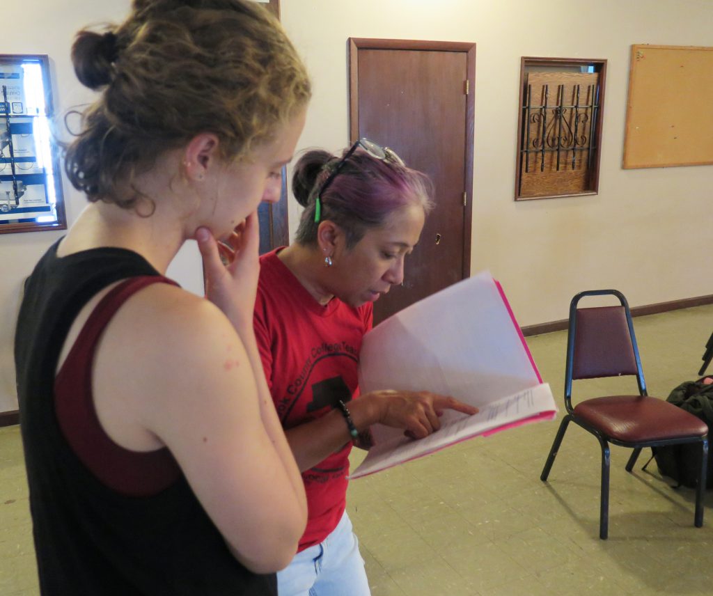 Image: Maxine Patronik (left) and Lani T. Montreal (right) at a rehearsal for their Body Passages piece at the Chicago Danztheatre Ensemble Auditorium. The artists stand next to each other, right sides in profile, looking down at a piece of paper in a folder in Lani’s hand. Lani points toward the center of the page as Maxine looks on, right hand to her face. Lani wears a red t-shirt and light jeans. Maxine wears a black tank-top with a maroon top underneath. Covered windows, a door, a bulletin board, and chairs are in the background. Photo by Marya Spont-Lemus.