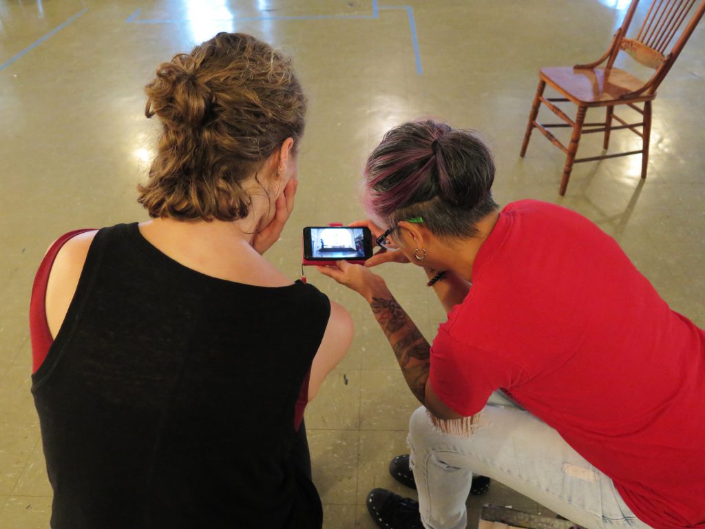 Image: Maxine Patronik and Lani T. Montreal at a rehearsal at Chicago Danztheatre. The artists sit side-by-side in chairs, backs toward the camera. Lani holds a camera-phone between them as they watch its bright screen, which shows a recording of themselves performing in the same space. Lani wears a red t-shirt and light jeans. Maxine wears a black tank-top with a maroon strap beneath. The auditorium’s floor and a chair are in the background. Photo by Marya Spont-Lemus.