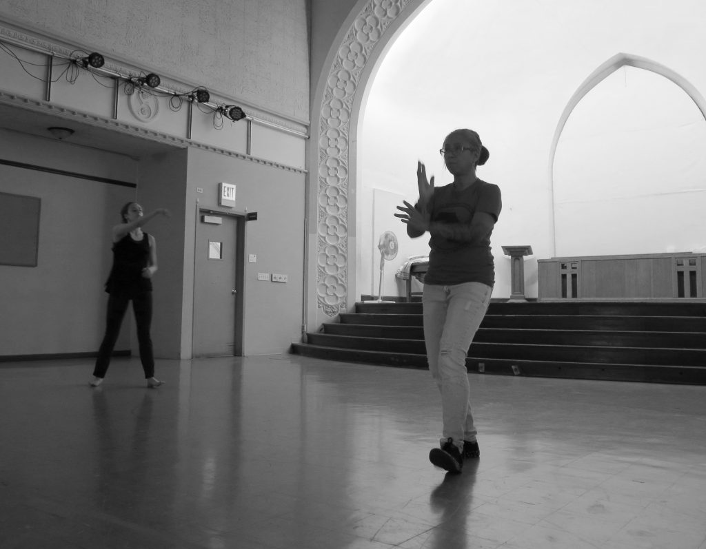 Image: Maxine Patronik and Lani T. Montreal rehearse for their Body Passages piece at the Chicago Danztheatre Ensemble Auditorium. In this black-and-white image, Maxine and Lani stand upright on the floor in front of the stage, roughly perpendicular to each other and both facing diagonally past the camera. In the foreground, Lani raises her arms in front of her chest, perpendicular to each other and with palms open, and leans on her back foot. In the background, Maxine lifts one arm straight ahead of her and holds the other by her abdomen. Lani wears a dark t-shirt and light jeans. Maxine wears a dark tank-top and dark pants. Photo by Marya Spont-Lemus.