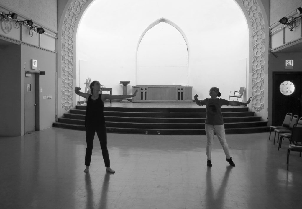 Image: Maxine Patronik and Lani T. Montreal rehearse for their Body Passages piece at the Chicago Danztheatre Ensemble Auditorium. In this black-and-white image, both artists stand dark in the middle of the floor, with the stage bright in the background. Maxine’s and Lani’s bodies face the camera, as the artists face each other. They strike a similar pose — feet spread shoulder-width apart, left arms stretching to their left sides, left wrists cocked, and right arms bent to their right sides. Lani wears a dark t-shirt, light jeans, and shoes. Maxine wears a dark tank-top and dark pants and is barefoot. Photo by Marya Spont-Lemus.