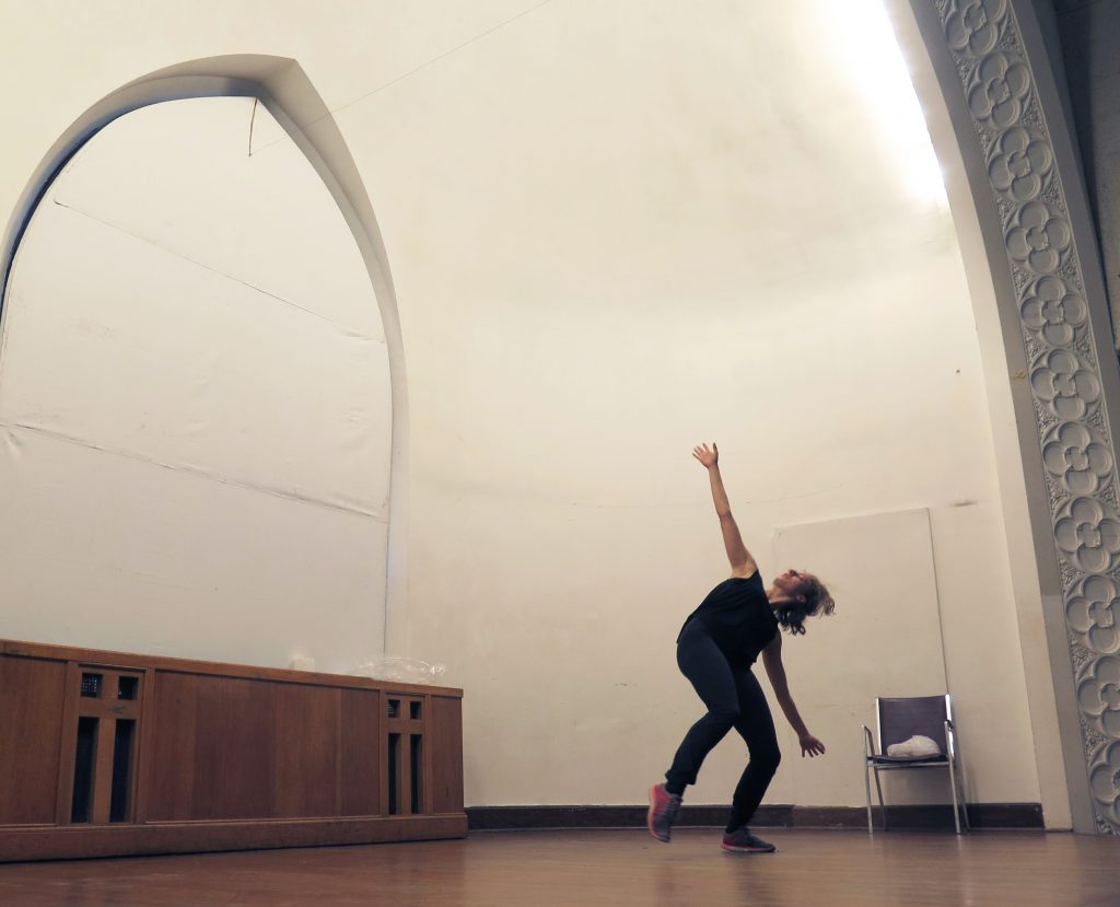 Image: Maxine Patronik rehearses on-stage at Chicago Danztheatre. Her body faces front. Maxine appears mid-motion, leaning her body far to her left, stretching her arms toward the floor and the ceiling, and looking up at the ceiling. She balances on her left foot, with her right foot pointed and off the floor. Both knees are slightly bent. Maxine wears a black tank-top and black pants. Photo by Marya Spont-Lemus.