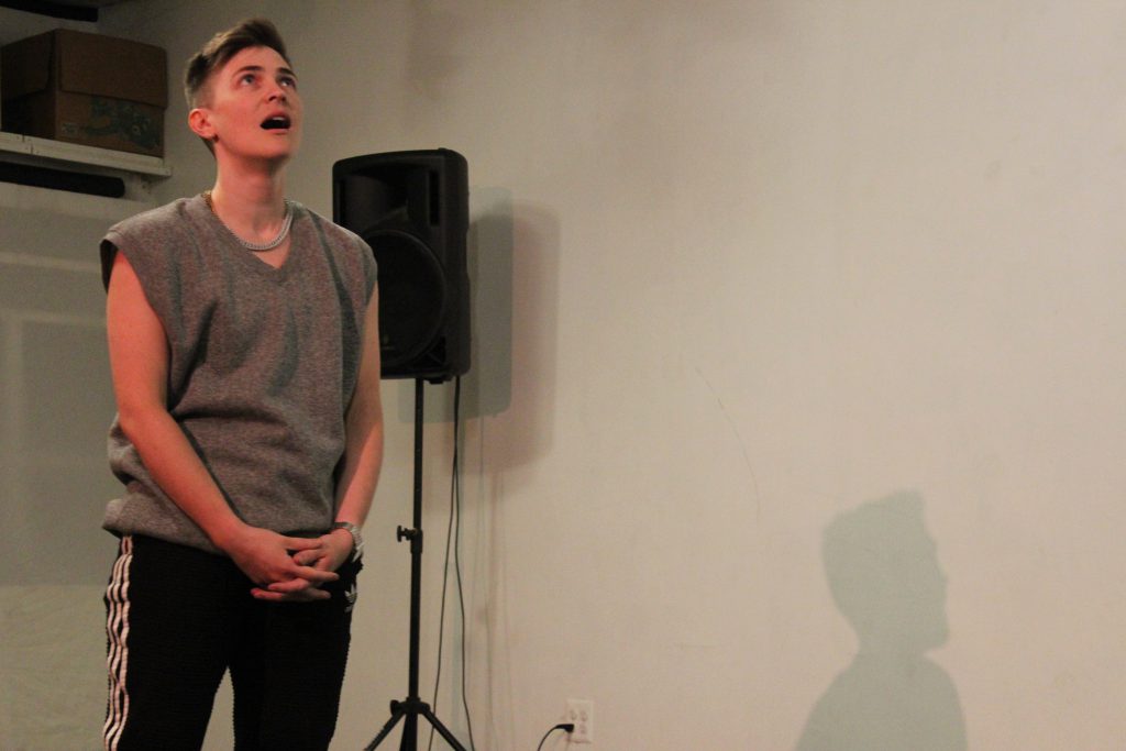 Image: Nora Sharp performs in response to the score “Dear Aron: (Destroy) Armor.” On the left side of the image, the performer stands with hands folded, looking up at the ceiling and singing. On the right side, part of Nora’s shadow is visible, cast on the wall. The artist wears a grey sleeveless shirt and black athletic pants with white stripes. Photo by Caleb Neubauer.