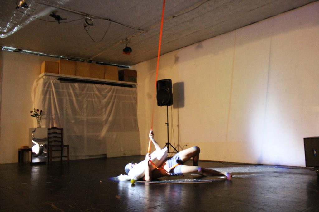 Image: Regin Igloria performs in response to the score “Dear Regin: SOAK (in silence).” Regin lies on the floor of TriTriangle, lit by a spotlight. Regin is barefoot and wears a swim cap, goggles, and spandex shorts. A sandbag lies across Regin’s chest. The artist reaches his arms around the sandbag to grip an orange strap that hangs from the ceiling, as he pushes his hips off the floor. Photo by Caleb Neubauer.